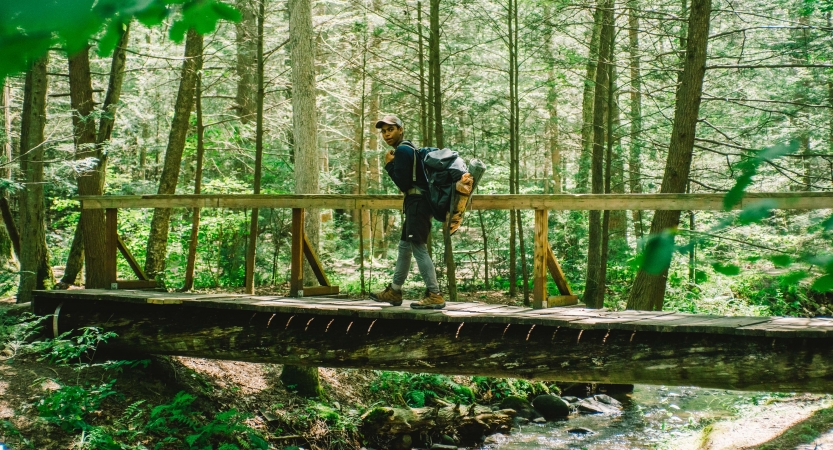 a young person carrying a backpack crosses a wooden bridge over a creek in a wooded area